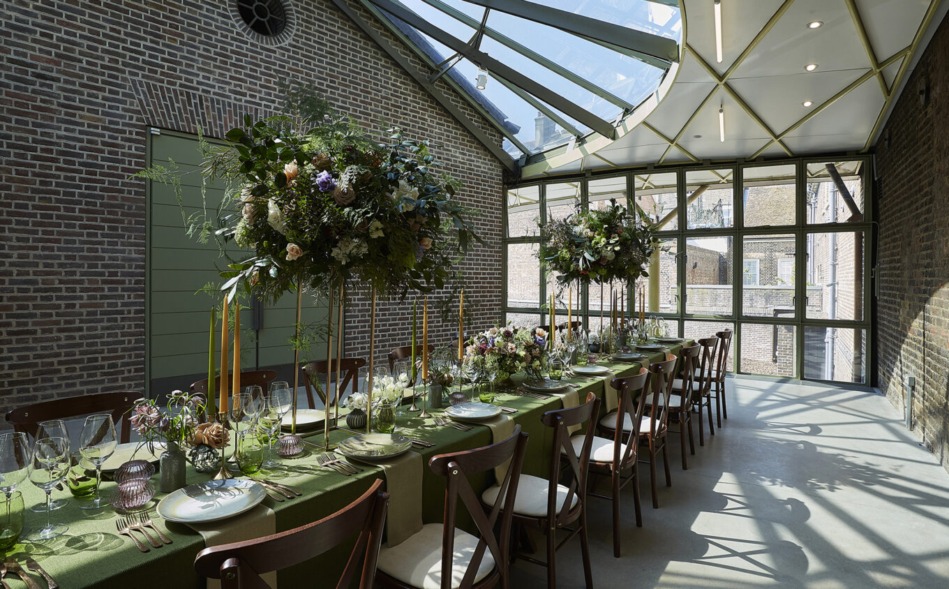A long dining table dressed with greenery in a glass atrium