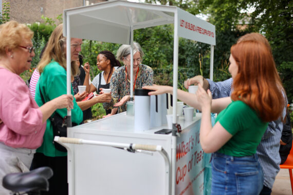 A group of people gather around a mobile tea cart outdoors. A woman serves tea while others chat and smile, holding paper cups. Trees and greenery provide a leafy backdrop.