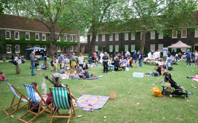 Large green lawn in front of historic brick buildings, with families sitting on blankets and deck chairs, children playing.