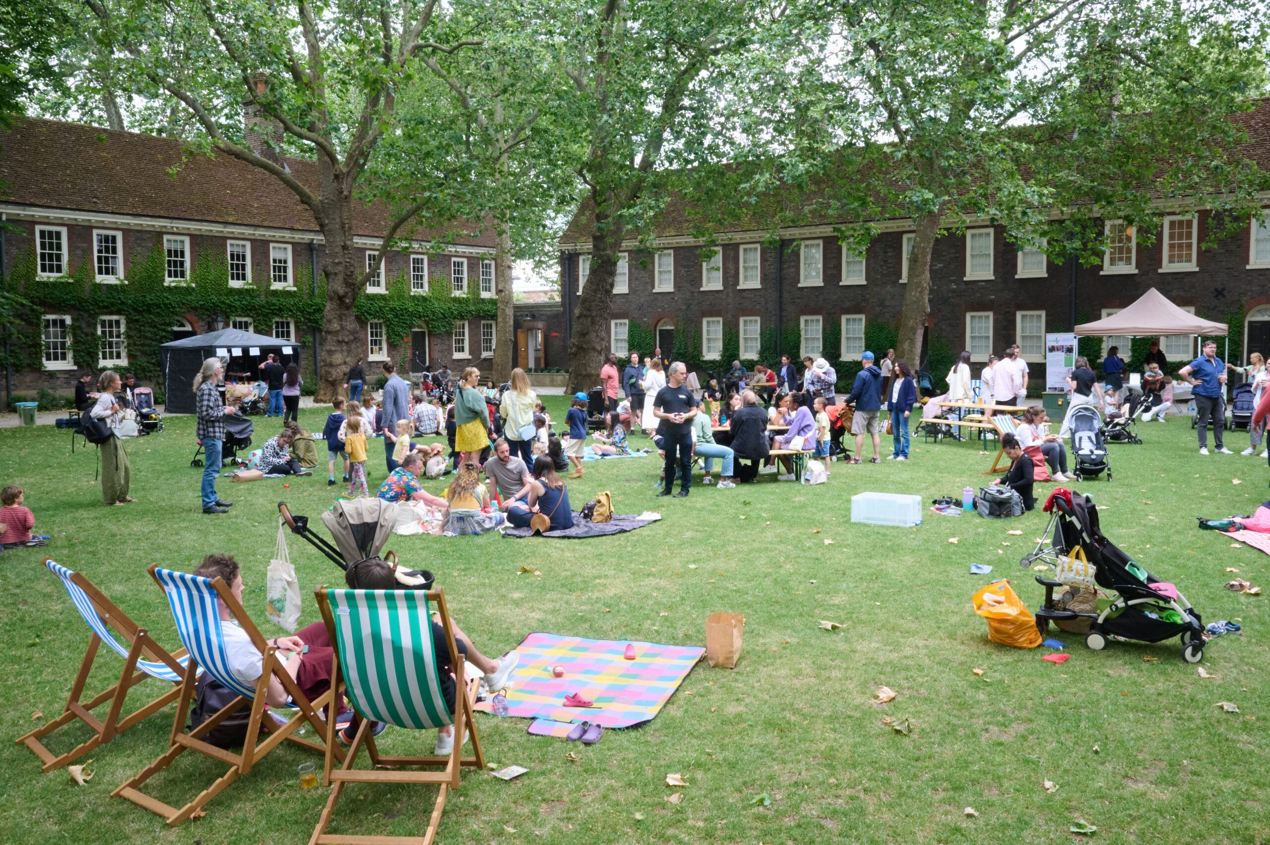 Large green lawn in front of historic brick buildings, with families sitting on blankets and deck chairs, children playing.