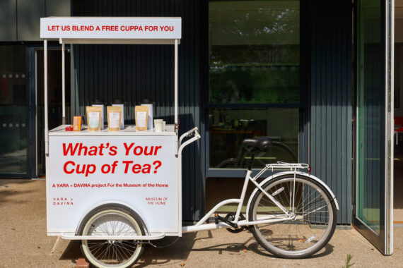 A white tricycle cart outside a building, with large red text reading “What’s Your Cup of Tea?”. Tea ingredients and takeaway cups are arranged on top under a canopy inviting free tea blending.