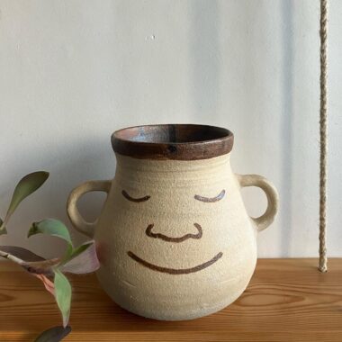 A beige ceramic mug with a wide base and two handles, featuring a painted smiling face with closed eyes. It sits on a wooden surface next to a leaf.