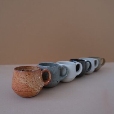 A line-up of six ceramic espresso cups in a range of matte glazes—spotted terracotta, grey, off-white, black, cream, and brown—arranged on a pinkish grey surface against a brownish beige background.