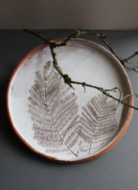 A round ceramic plate viewed from above, with a terracotta rim and white glazed surface, adorned with delicate brown fern leaf prints. A mossy branch rests across the plate.