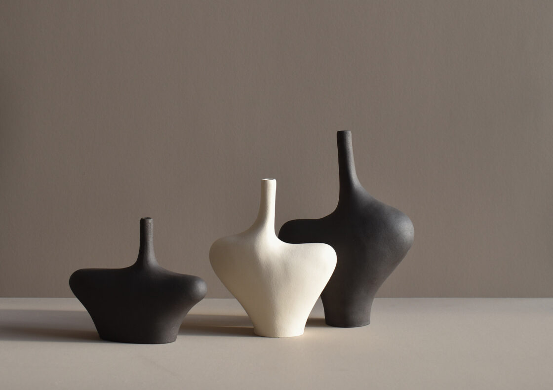 Three matte ceramic vases with abstract forms. Two are charcoal black and one is off-white, arranged side by side against a neutral background with soft shadowing.