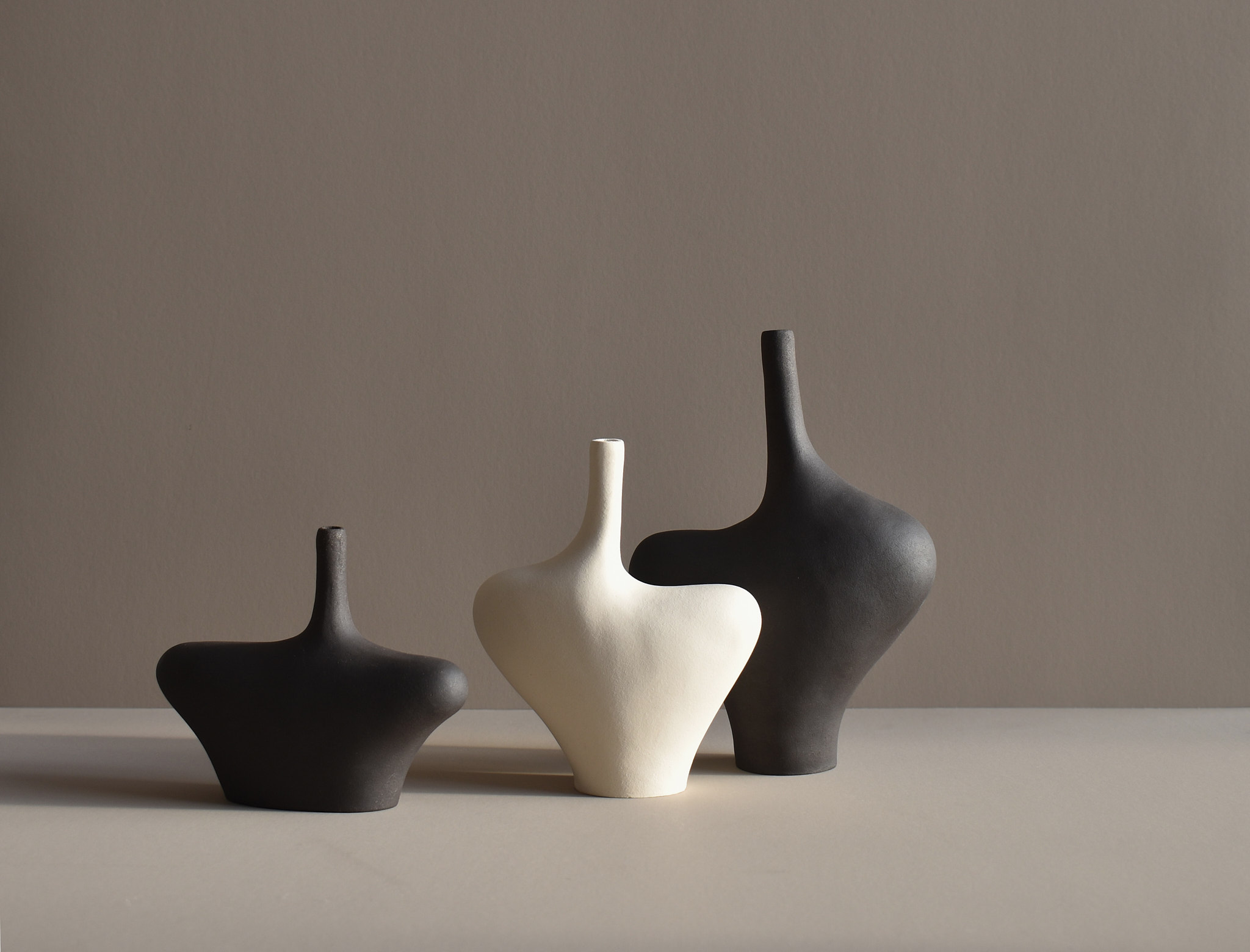 Three matte ceramic vases with abstract forms. Two are charcoal black and one is off-white, arranged side by side against a neutral background with soft shadowing.