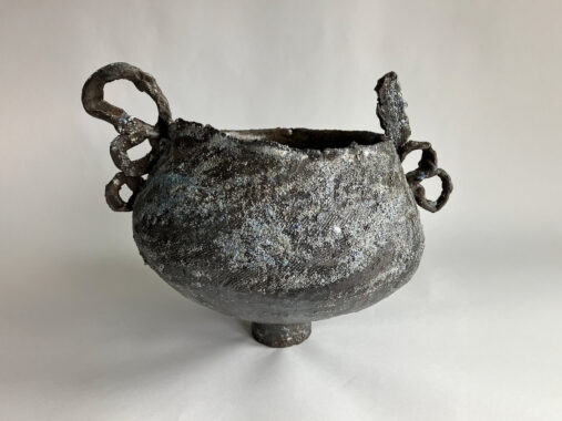 A heavily textured, dark brownish grey ceramic vessel with a rounded body, small base, and looping sculptural handles. Its rough surface appears encrusted, giving it a volcanic, ancient look.