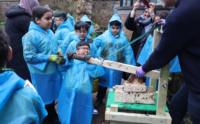 Children in blue rain ponchos outside in a garden making adobe bricks and smiling