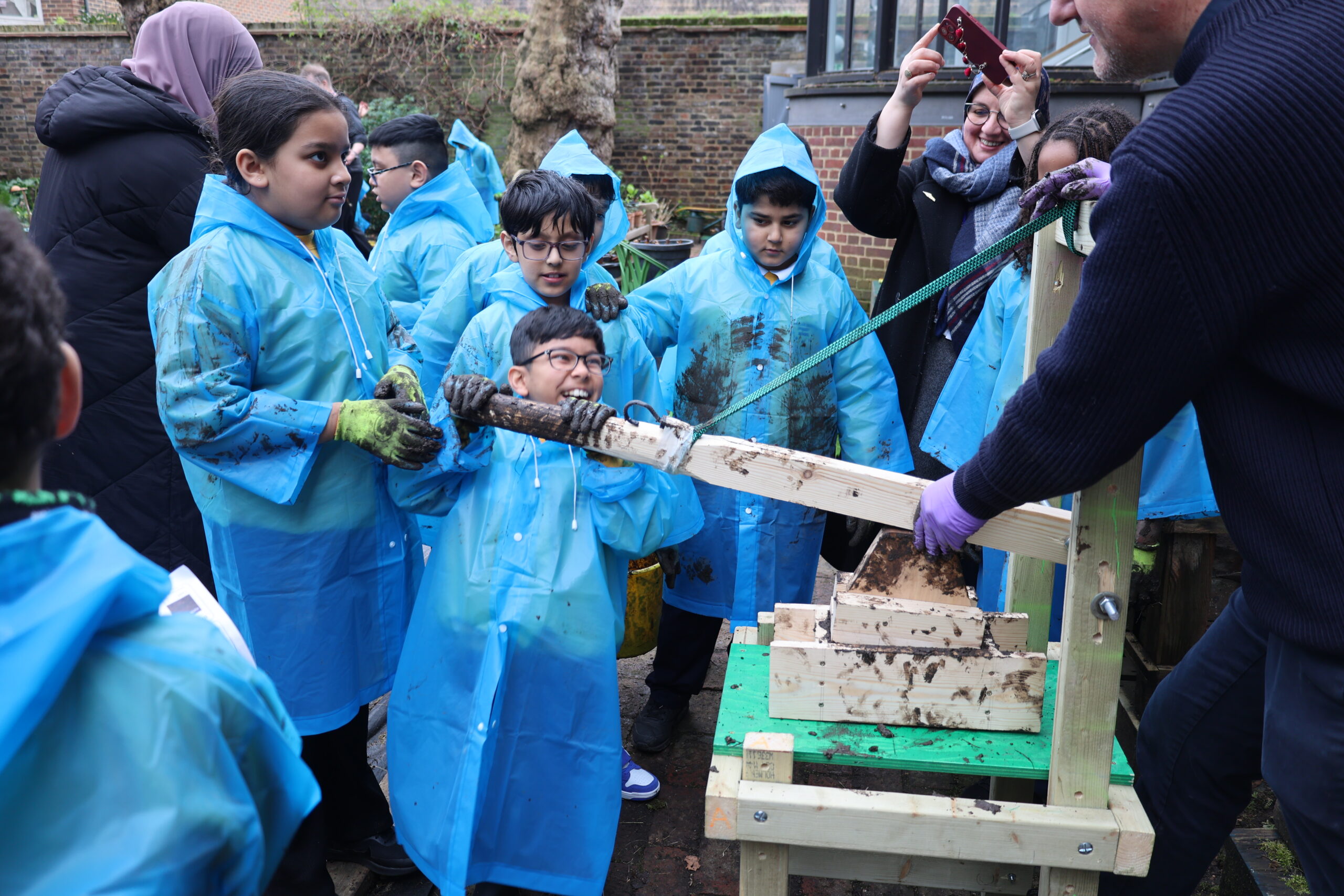 Children in blue rain ponchos outside in a garden making adobe bricks and smiling
