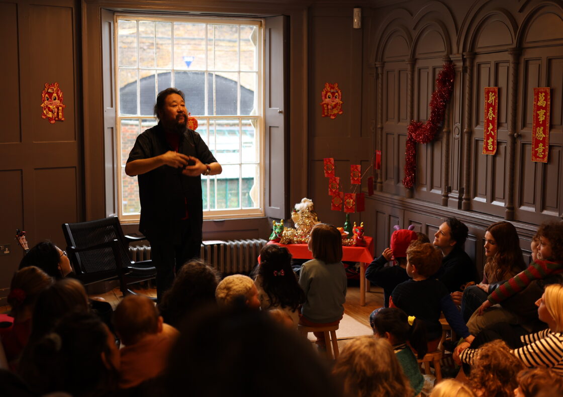 Children sit on stools watching a man motion to them.