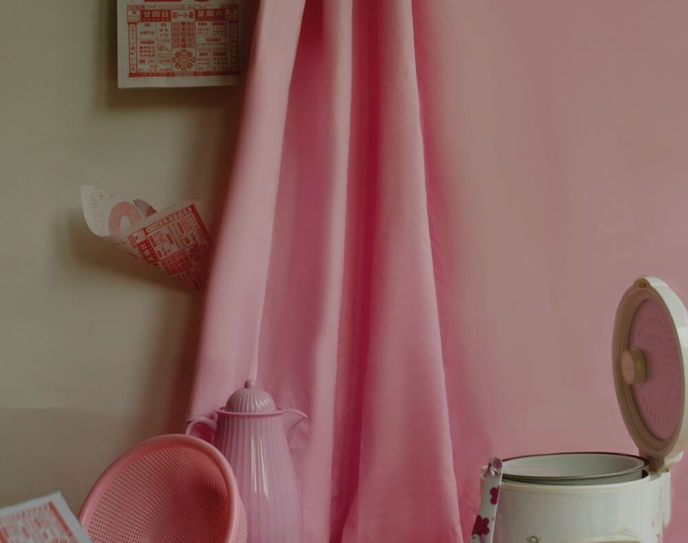A still life with household items including a rice cooker, colander, teapot, and calendar against pink a curtain.
