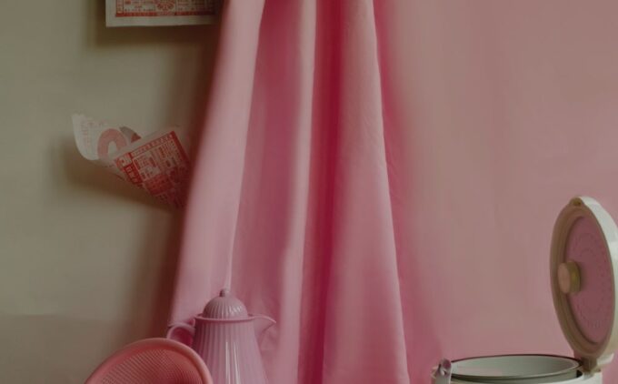 A still life with household items including a rice cooker, colander, teapot, and calendar against pink a curtain.