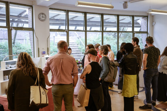 A crowd of people browsing and shopping at an indoor ceramics fair, with display tables visible and large windows in the background.