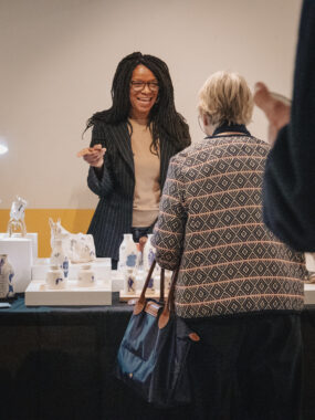 Two women having a conversation at a ceramics exhibition, with ceramics displayed on a table between them.