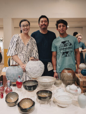Three people posing behind a table displaying various ceramic pottery pieces including bowls, vases, and decorative items.