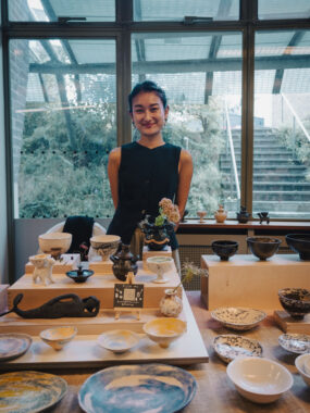 A young woman in a black sleeveless top standing behind a table displaying various ceramic bowls, plates, and pottery items near large windows.