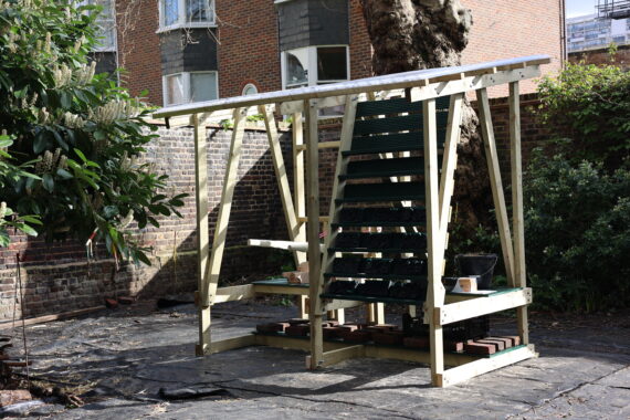 A wooden-framed structure with shelves and a roof structure stands in a corner around a brick wall and leafy plants in a garden.