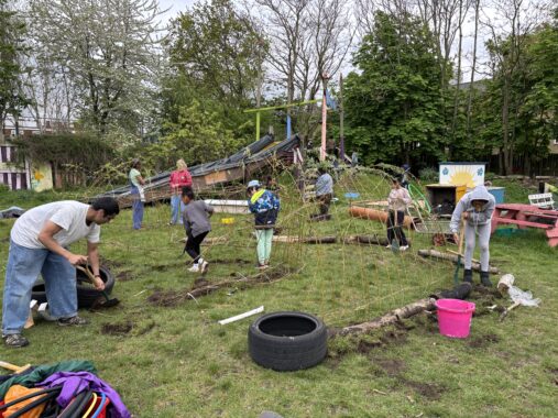 A group of adults and children bend willow branches into a dome-like shape, forming the structure of a living shelter on a grassy outdoor site.