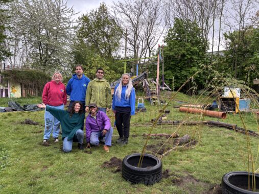 A smiling group of seven adults in colourful hoodies pose in front of a willow dome structure in a vibrant, community-run green space.