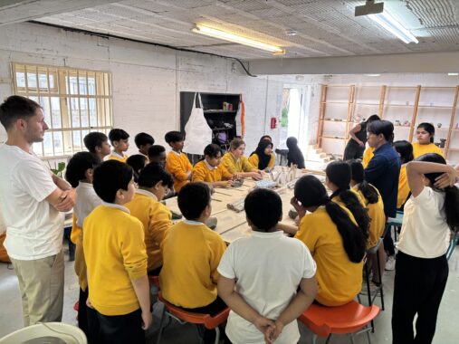 A class of schoolchildren in yellow and white shirts gather around a long table in a ceramics workshop, listening to the instructor.