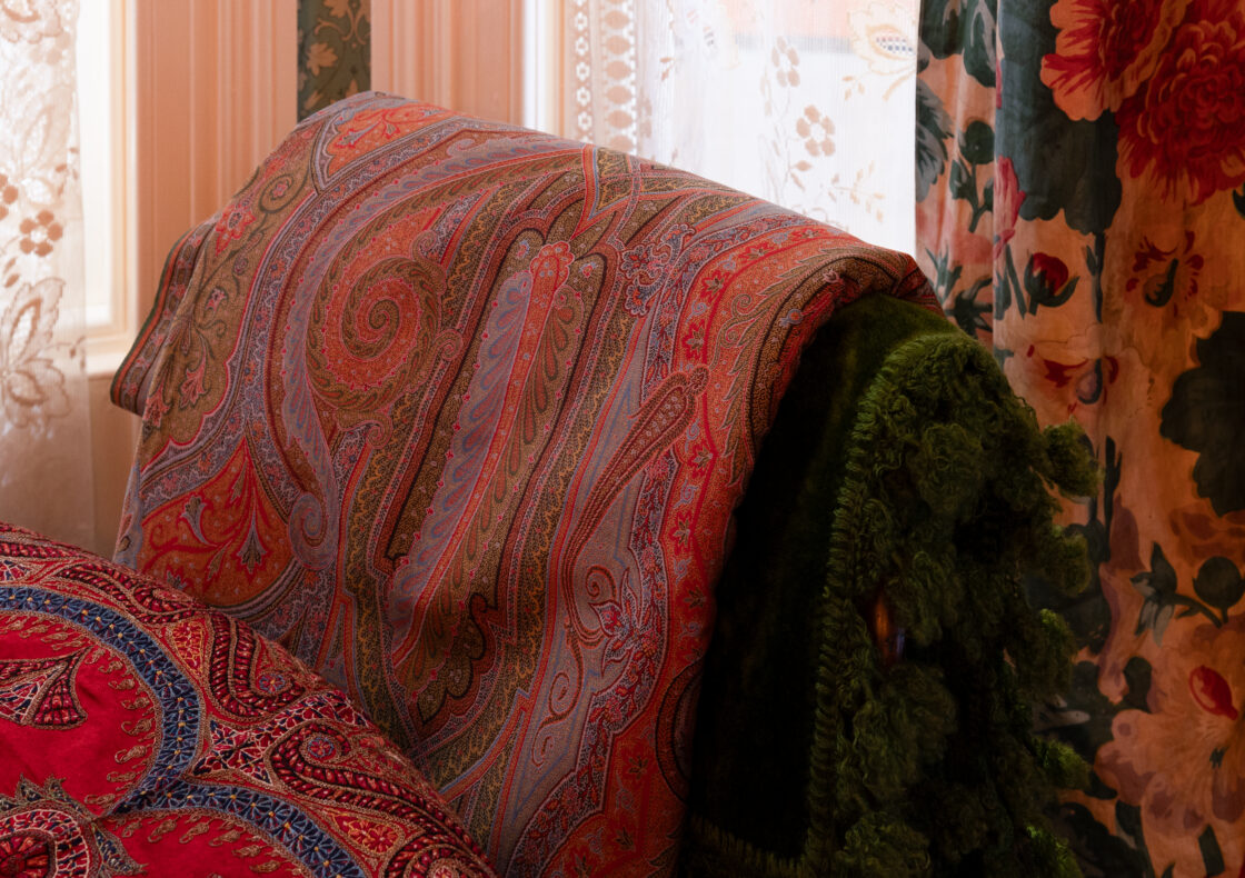 Red paisley-patterned pashmina draped over green velvet chair against floral curtains.