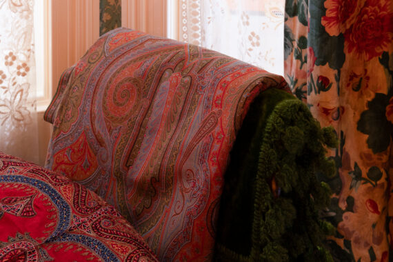 Red paisley-patterned pashmina draped over green velvet chair against floral curtains.