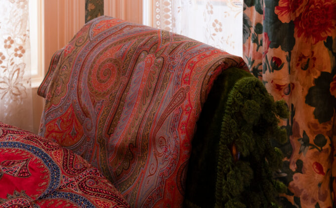 Red paisley-patterned pashmina draped over green velvet chair against floral curtains.