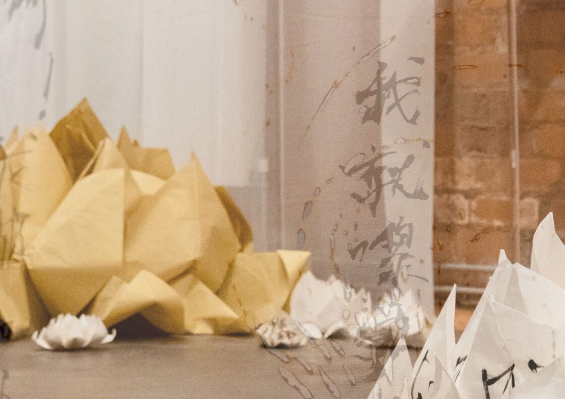 A close up installation view of artworks featuring a large folded paper lotus forms in warm yellow and smaller ones in ivory, scattered across a smooth floor. The image is overlaid with a graphic of calligraphic fonts.
