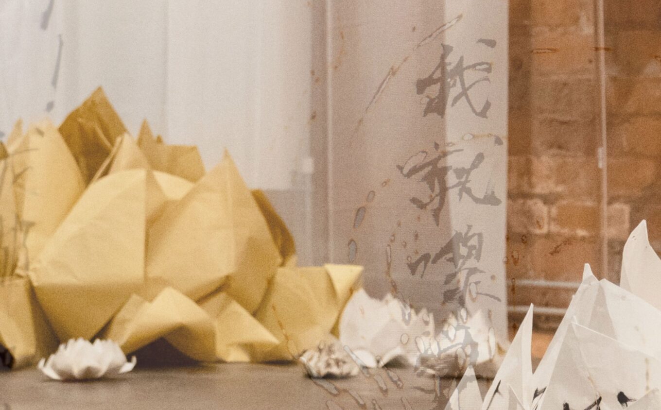 A close up installation view of artworks featuring a large folded paper lotus forms in warm yellow and smaller ones in ivory, scattered across a smooth floor. The image is overlaid with a graphic of calligraphic fonts.