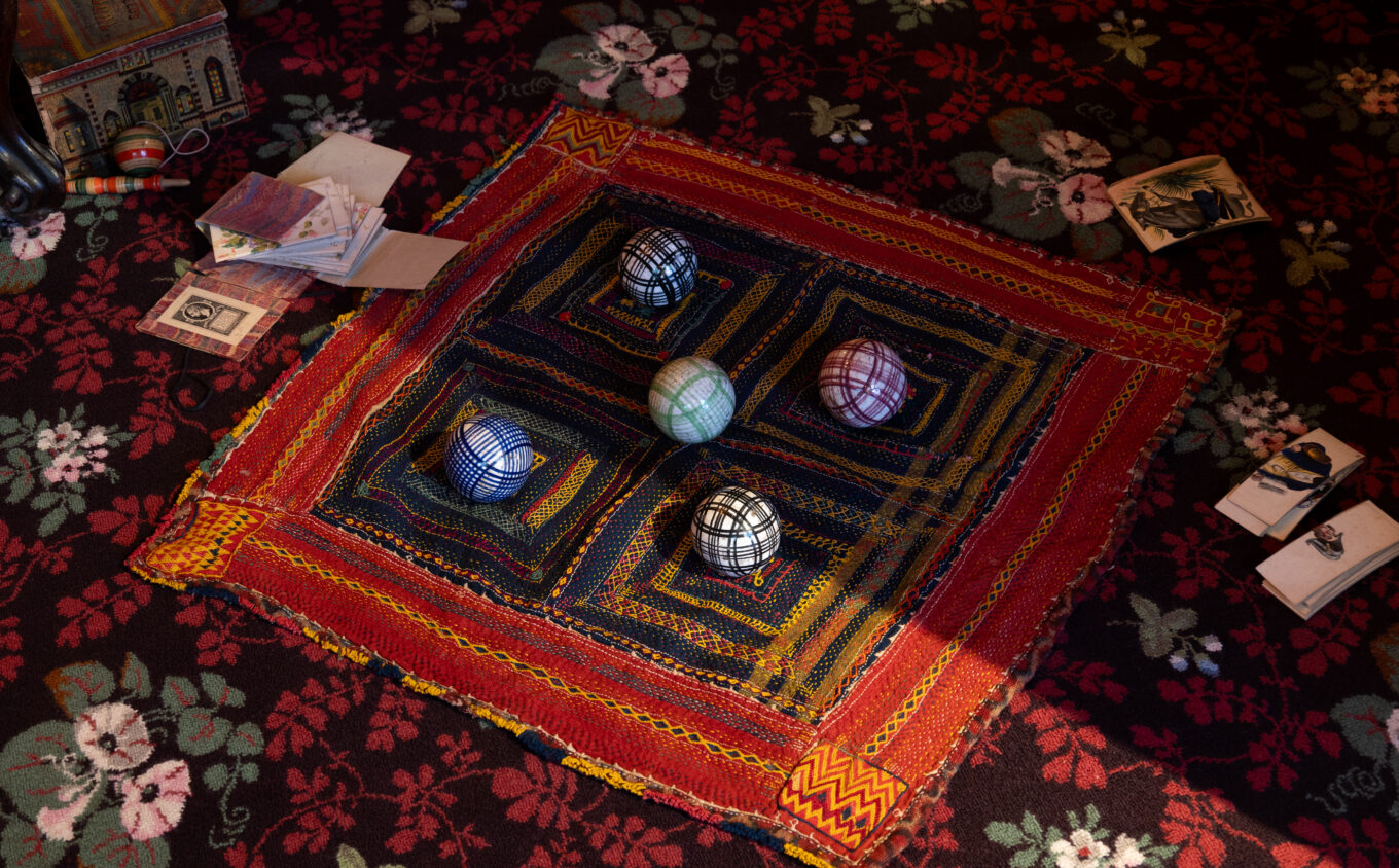 A richly patterned red, black, and floral carpet with a stitched square textile laid at the centre, bordered in red, yellow, and orange. Five colourful check-patterned balls sit atop the textile. Scattered around the edges are old-fashioned toys, paper cards, and a small illustrated box, suggesting a Victorian-era play setting. The scene is lit with dim but warm light, with shadows cast diagonally across the composition.