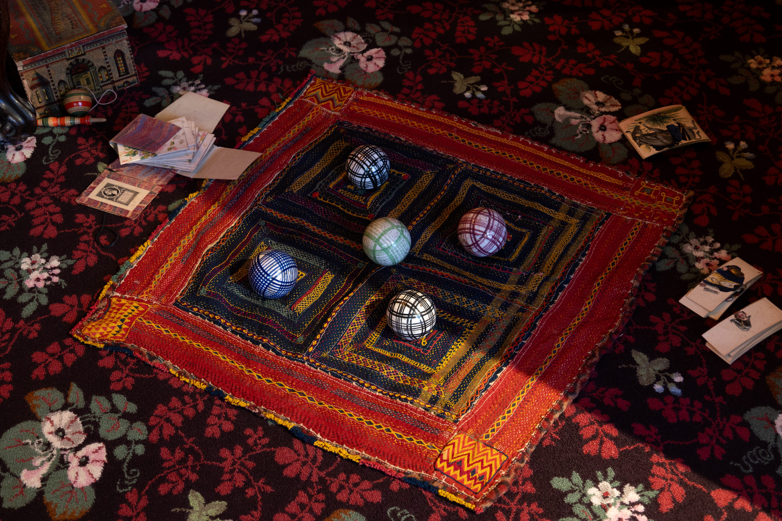 A richly patterned red, black, and floral carpet with a stitched square textile laid at the centre, bordered in red, yellow, and orange. Five colourful check-patterned balls sit atop the textile. Scattered around the edges are old-fashioned toys, paper cards, and a small illustrated box, suggesting a Victorian-era play setting. The scene is lit with dim but warm light, with shadows cast diagonally across the composition.