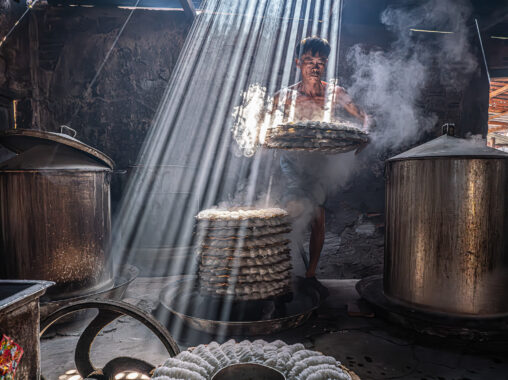 In a steamy kitchen, rays of sunlight pierce through the roof, illuminating stacks of delicate rice cakes as a person carefully handles them amidst towering metal vats.