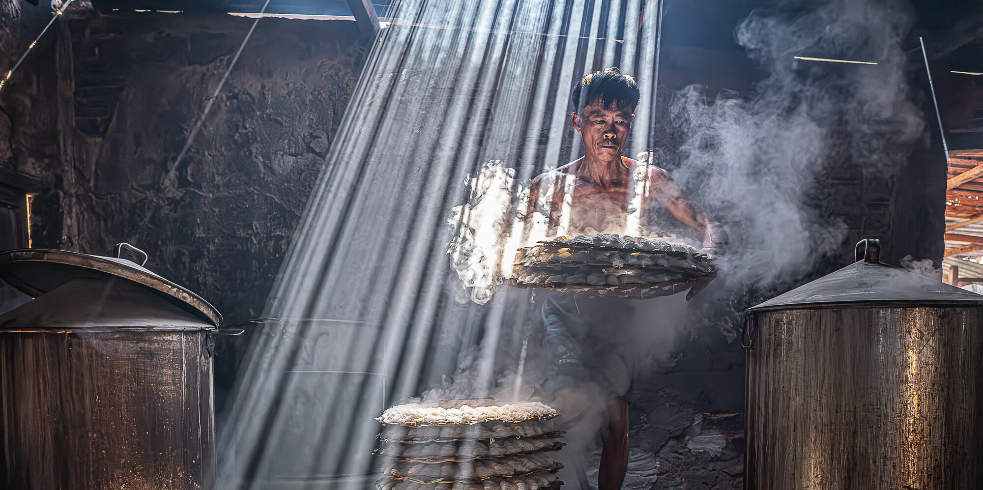 In a steamy kitchen, rays of sunlight pierce through the roof, illuminating stacks of delicate rice cakes as a person carefully handles them amidst towering metal vats.