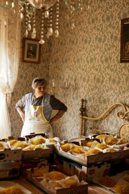 An elderly woman in a patterned apron stands in a softly lit, vintage-styled bedroom, hands on hips, surveying trays of golden homemade pasta laid out on a bed and table.