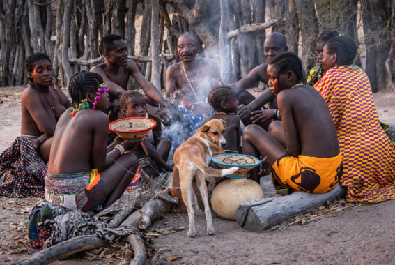A circle of people in thin, vibrant, patterned clothes gathered on sandy ground, one of them holding a bowl. A calm dog stands among them. Smoke rises gently from the middle of the group (likely from a fire made with gathered tree branches, though the flames are hidden from view). The smoke drifts upward and outward, partially veiling the bare tree trunks and rustic wooden fence in the background.
