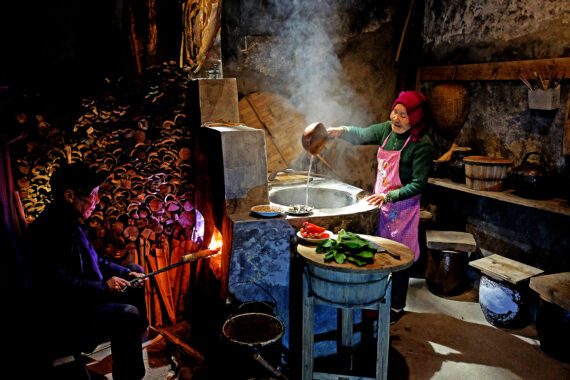 In a rustic, dimly lit kitchen, an elderly person pours water from a ladle into a traditional stone stove as steam rises into the air. Nearby, an elderly person stokes the fire with glowing embers. Around them are piles of chopped wood, vegetables on a wooden table, and kitchen tools.