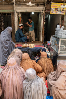 A group of women sit and wait in front of a market as they wait for free bread.