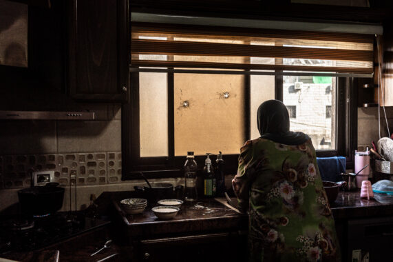 A woman in a floral robe and hijab leans by a kitchen sink, facing a cracked window pierced by bullet holes.