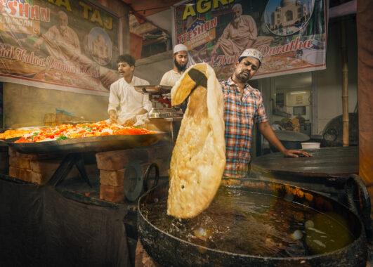 Inside a food stall, a person in a checkered shirt stands in the foreground, mid-motion as he lifts an enormous golden doughy flatbread into a sizzling deep fryer. In the back are two people in white clothes, working on food.