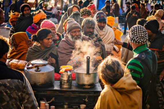 A crowd wrapped in shawls and winter layers queues closely around a steaming street food stall. The golden sunlight shines on them.