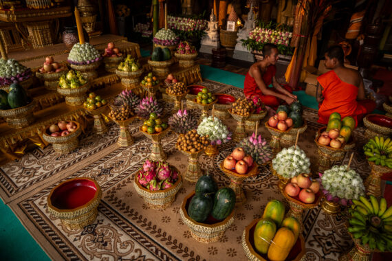 Inside a temple, two monks in dark orange robes sit in quiet conversation, surrounded by an elaborate spread of fruits, such as dragon fruits and apples, and flowers arranged on ornate golden pedestal trays. These trays are carefully placed on richly patterned carpets that add warmth and detail to the setting. In the background, Buddhist statues and floral offerings line the altar.