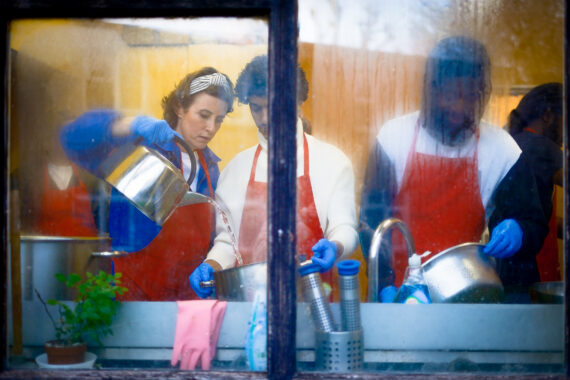 Three people in red aprons and blue latex gloves are washing pans by the sink, viewed through a misted window. The person on the left pours water from a metal pot into another that the person in the middle is holding. The person on the right is looking down at the pot he is holding.
