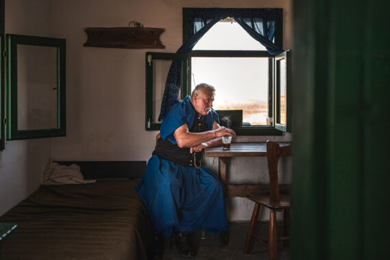 An older man dressed in blue garments sitting by the window in a mall, tightly arranged room. He leans one arm on a wooden table beside a steaming glass. window, framed by dark green shutters and a patterned blue curtain.