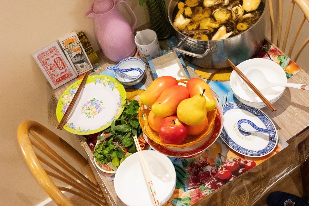 A table with dishes, fruit in a basket, cooking pot, and various dining items spread across the surface.