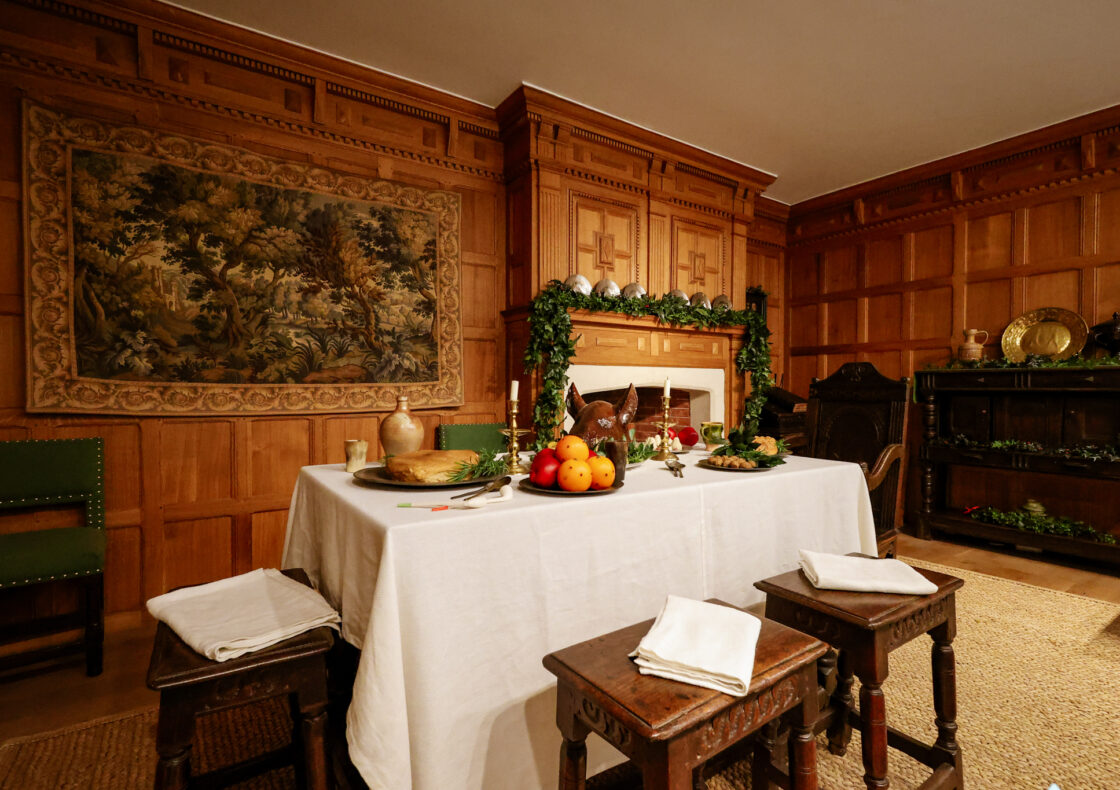 A table is laid with fruit, candles, greenery and a pigs head, in a room with wood paneling, a fireplace and a tapestry.