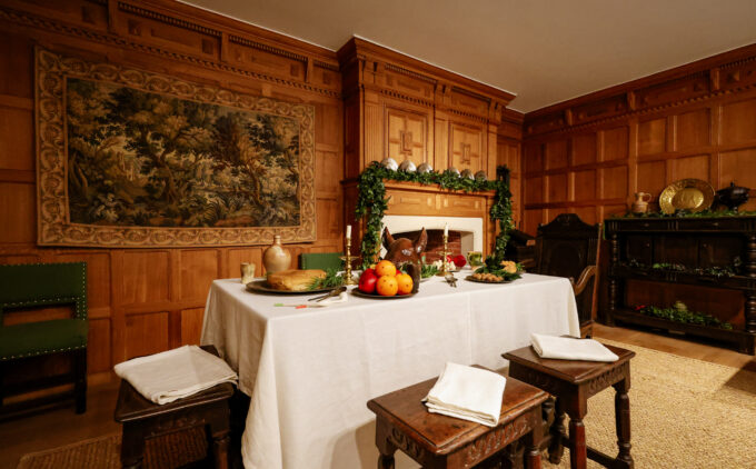 A table is laid with fruit, candles, greenery and a pigs head, in a room with wood paneling, a fireplace and a tapestry.