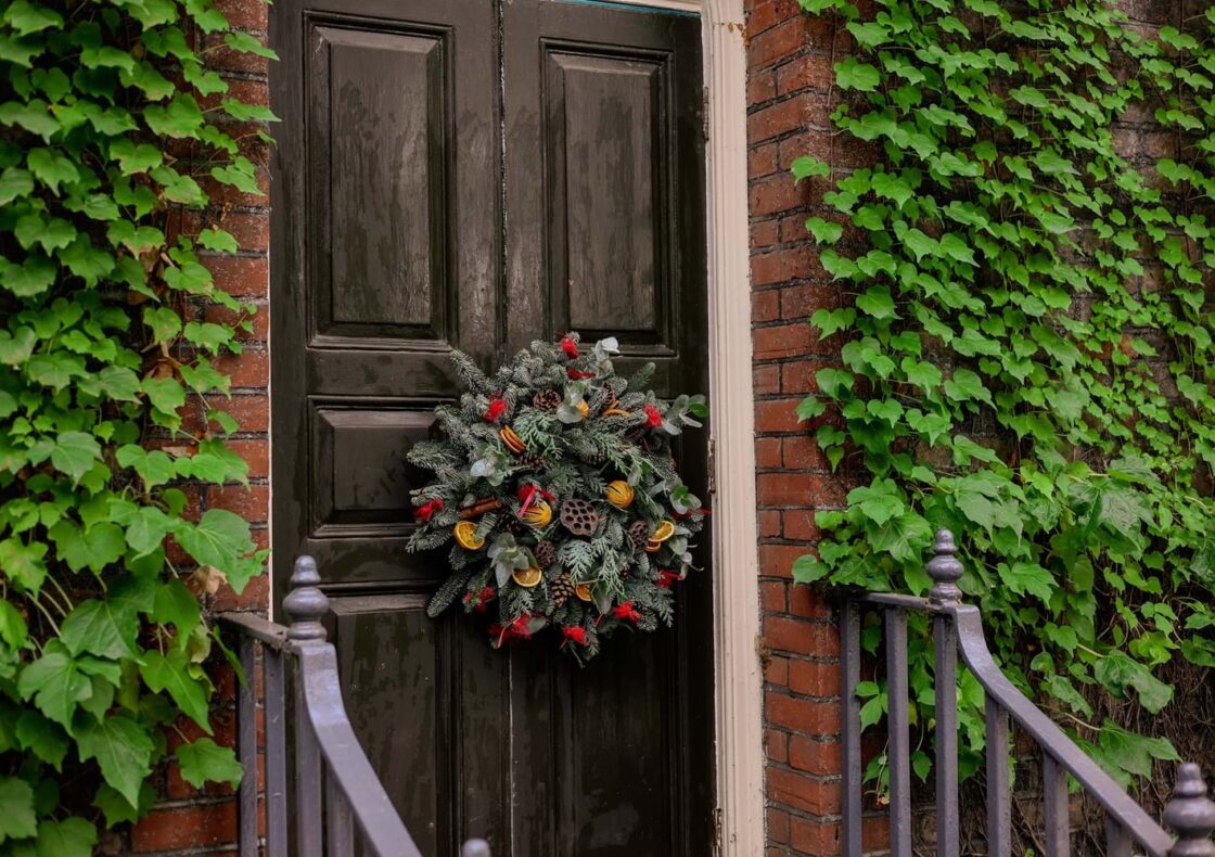 A decorative wreath with dried oranges and red berries adorns a wooden door, with ivy covered walls.