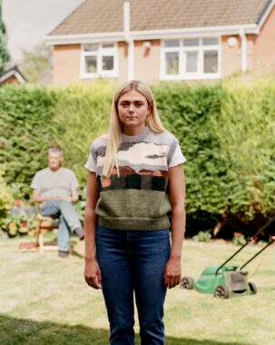 Young blonde woman in landscape-patterned knit vest standing in suburban backyard.