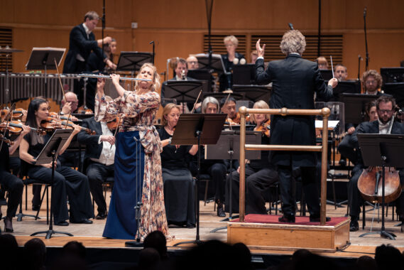 Orchestra performance with female flutist soloist in colourful dress and conductor on concert hall stage.