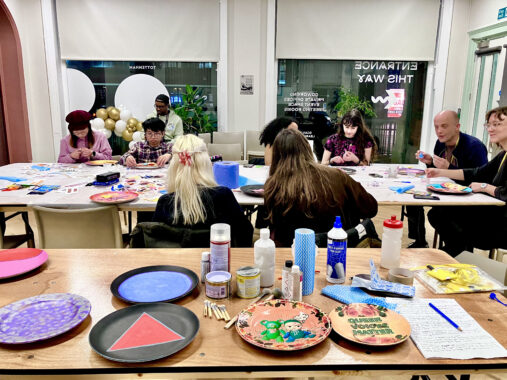 Group workshop with people seated around tables covered in craft supplies and materials.